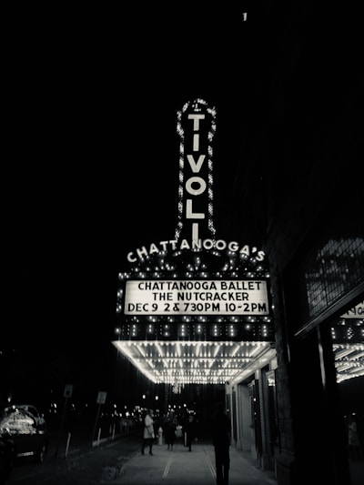 A black and white photograph of a theater entrance with a marquee that reads 'CHATTANOOGA BALLET THE NUTCRACKER DEC 9 2 & 7:30PM 10-2PM' in illuminated letters. The large signage of the Tivoli Theatre is vertical and well-lit. People walk underneath the marquee on the sidewalk, and the street is lined with lights and signs.