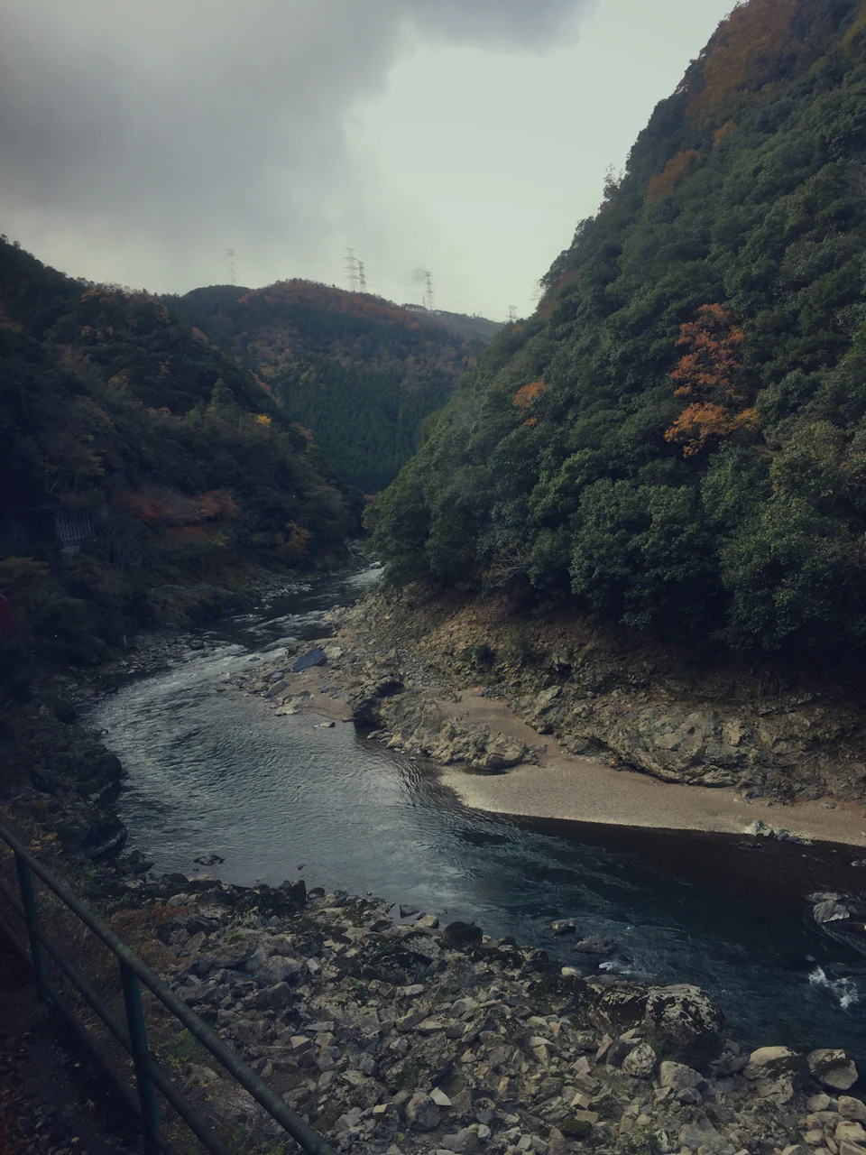 The image depicts a serene mountain landscape with a river flowing through it, surrounded by lush green trees and shrubs on both sides. The sky above is overcast, casting a soft light over the scene. In the foreground, there are rocks and boulders scattered along the riverbank, adding texture to the landscape.