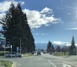 A scenic road with a mountain range in the background, bordered by tall trees and scattered clouds in a blue sky. Several cars are driving along the road, and there are road signs indicating directions. The landscape is open and spacious, evoking a sense of tranquility.