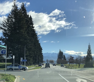 A scenic road with a mountain range in the background, bordered by tall trees and scattered clouds in a blue sky. Several cars are driving along the road, and there are road signs indicating directions. The landscape is open and spacious, evoking a sense of tranquility.