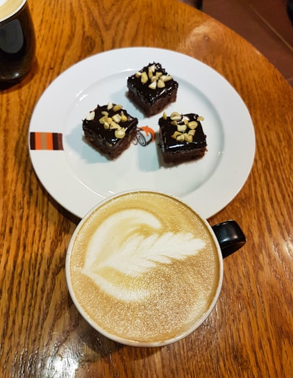 A plate with three small chocolate cakes topped with chopped nuts. Next to the plate is a cup of cappuccino with intricate latte art on a wooden tabletop.