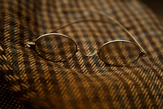 A pair of oval-shaped eyeglasses with thin metal frames is resting on a textured, patterned fabric. The fabric has alternating diagonal stripes in warm earth tones, creating a cozy and vintage feel.