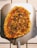 A freshly baked pizza placed on a metallic pizza peel. The pizza has a thin, crispy crust and is topped with melted cheese, small diced green peppers, and caramelized edges. The background consists of white tiled flooring with brown grout lines.