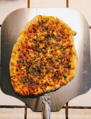 A freshly baked pizza placed on a metallic pizza peel. The pizza has a thin, crispy crust and is topped with melted cheese, small diced green peppers, and caramelized edges. The background consists of white tiled flooring with brown grout lines.