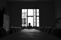 A person sits alone at the far end of a long conference table in a dimly lit room. The window behind them is open, allowing light to stream in and silhouette the individual. The room has minimal decor with some patterned artwork on one wall and a series of small images or notes on another. The chairs around the table are empty, emphasizing solitude.