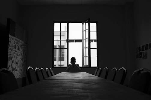 A person sits alone at the far end of a long conference table in a dimly lit room. The window behind them is open, allowing light to stream in and silhouette the individual. The room has minimal decor with some patterned artwork on one wall and a series of small images or notes on another. The chairs around the table are empty, emphasizing solitude.