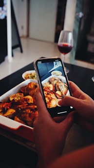 Hands holding a smartphone capturing a photo of a roasted chicken dish on a table. A glass of red wine and a bowl with other food are also present nearby, indicating a dining setting.