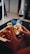 Hands holding a smartphone capturing a photo of a roasted chicken dish on a table. A glass of red wine and a bowl with other food are also present nearby, indicating a dining setting.