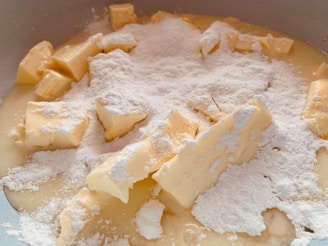 Chunks of butter are coated with a layer of fine white powdered sugar, mixing with a creamy liquid in a close-up view, suggesting the early stages of a baking preparation.