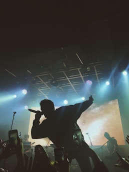 A silhouette of a singer holding a microphone on stage with bright spotlights and a large screen in the background. The audience is capturing the performance on their phones.