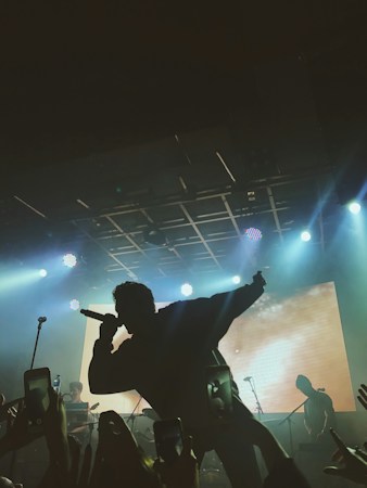 A silhouette of a singer holding a microphone on stage with bright spotlights and a large screen in the background. The audience is capturing the performance on their phones.