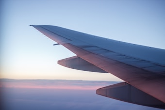 A photograph captures an airplane wing in flight against a serene sky. The wing extends diagonally across the image, with sunlight casting a warm glow along its edge. Soft layers of clouds and a gradient from soft pink to blue form the backdrop.