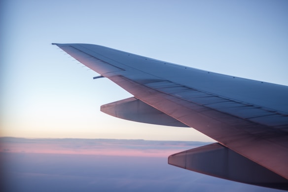 A photograph captures an airplane wing in flight against a serene sky. The wing extends diagonally across the image, with sunlight casting a warm glow along its edge. Soft layers of clouds and a gradient from soft pink to blue form the backdrop.