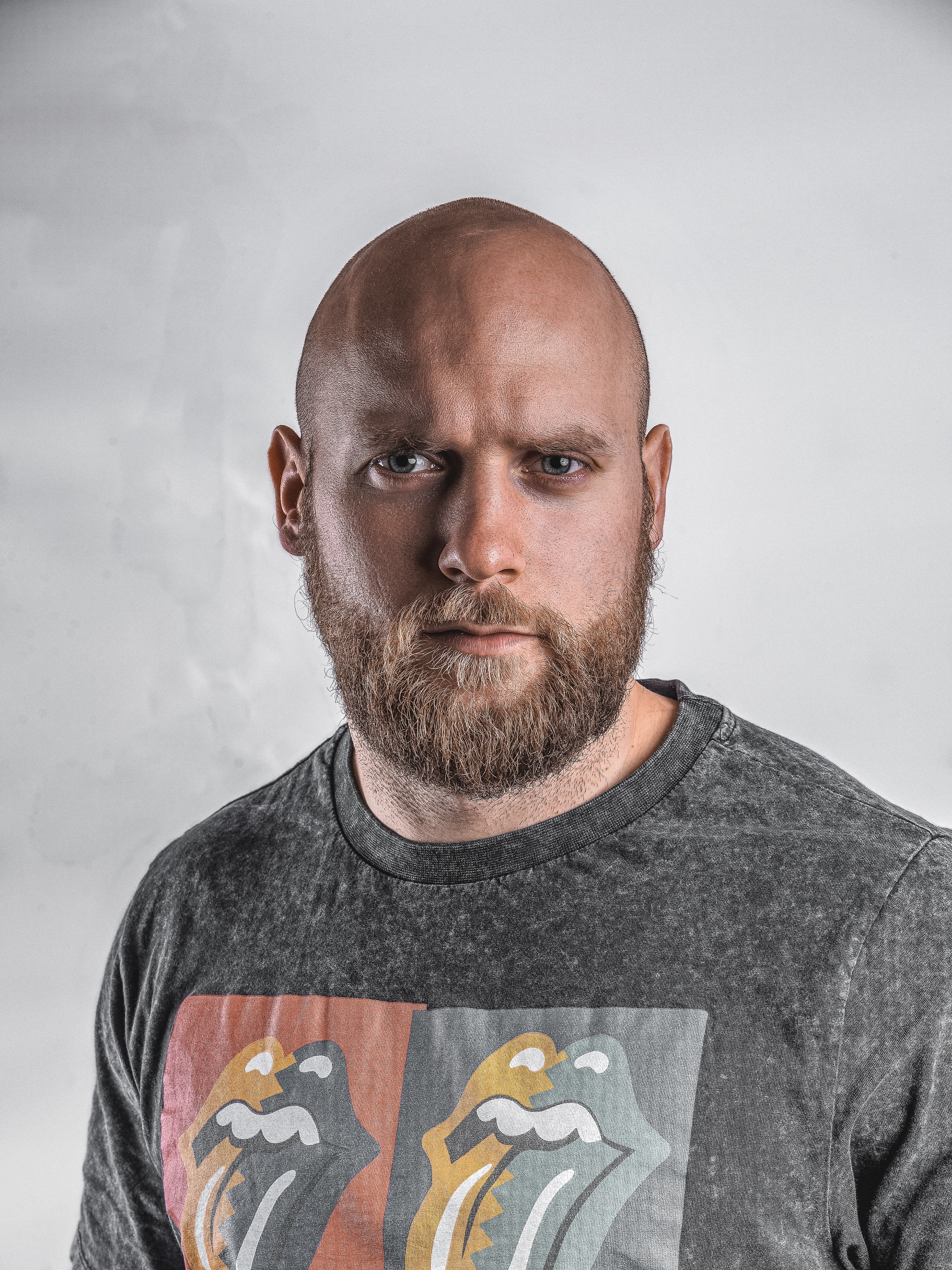 Bald man with a beard in a grey crew-neck top, neutral expression, in front of a light wall