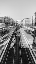 The image displays a train station with multiple tracks running through the center. Buildings line both sides of the station area, and several canopies cover the platforms on each side. The station appears to be empty, with no people visibly present. The image is in black and white, giving it a classic, somewhat nostalgic feel.