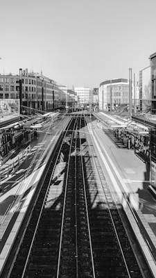 The image displays a train station with multiple tracks running through the center. Buildings line both sides of the station area, and several canopies cover the platforms on each side. The station appears to be empty, with no people visibly present. The image is in black and white, giving it a classic, somewhat nostalgic feel.