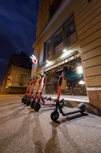 Several electric scooters are lined up on a paved sidewalk outside a building with a beige exterior. Above the scooters, a shop with a decorative sign and a heart-shaped neon light is visible. The scene is captured during evening or night, with artificial lights illuminating the area and casting shadows.