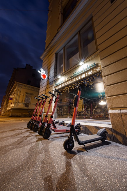 Several electric scooters are lined up on a paved sidewalk outside a building with a beige exterior. Above the scooters, a shop with a decorative sign and a heart-shaped neon light is visible. The scene is captured during evening or night, with artificial lights illuminating the area and casting shadows.