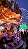 A lively amusement park scene with a carousel featuring a mix of small vehicles and horse figures. Bright lights illuminate the ride, creating a colorful display against the evening sky. A Ferris wheel is visible in the background while people gather around, enjoying the festive atmosphere.