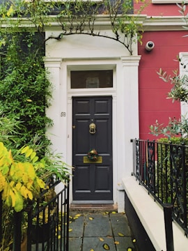 A dark grey door with a decorative wreath is set in a white-framed entryway. The building has a red exterior wall, and greenery, including a bush with yellow leaves, grows nearby. A security camera is mounted on the wall.