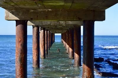 Perspective view under a pier showing rusty metal pillars extending into the ocean. The water is visibly blue, with small waves approaching. Sunlight casts shadows on the water and pillars, while the sky above is clear and bright.