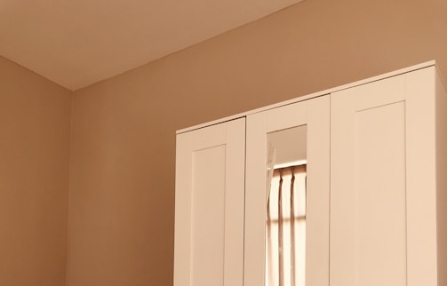 A section of a minimalist room featuring a white wardrobe against a light beige wall. The wardrobe includes a mirrored section that reflects part of a window with beige curtains. The clean lines and neutral tones create a calm, serene atmosphere.