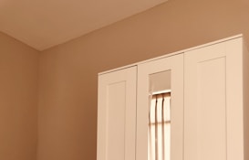 A section of a minimalist room featuring a white wardrobe against a light beige wall. The wardrobe includes a mirrored section that reflects part of a window with beige curtains. The clean lines and neutral tones create a calm, serene atmosphere.