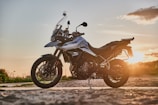 A sleek motorcycle is parked on a dirt road, set against the backdrop of a warm, golden sunset. The bike features a robust frame and metallic accents, suggesting a design focused on both performance and style. The surrounding landscape is open and tranquil, with hints of vegetation visible.