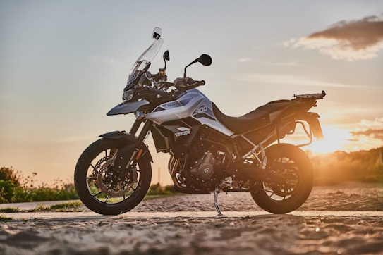 A sleek motorcycle is parked on a dirt road, set against the backdrop of a warm, golden sunset. The bike features a robust frame and metallic accents, suggesting a design focused on both performance and style. The surrounding landscape is open and tranquil, with hints of vegetation visible.