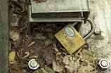 A metal lock is attached to a steel structure, surrounded by dried leaves and dirt. The lock is labeled with 'ABUS' and features text indicating rust resistance. There are bolts and a washer nearby on the ground.