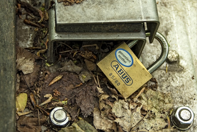 A metal lock is attached to a steel structure, surrounded by dried leaves and dirt. The lock is labeled with 'ABUS' and features text indicating rust resistance. There are bolts and a washer nearby on the ground.