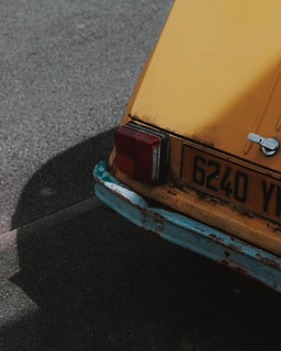 A close-up angle of an old, yellow car's rear end, showing visible rust and wear on its faded paint and bumper. The details include a metal latch, a tail light, and vehicle license plate with the number 6240 YL. The background is an asphalt road surface.