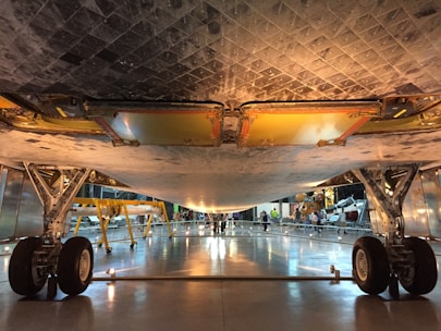 A view from below of a large, metallic aircraft or spacecraft with visible landing gear, structural elements, and tile-like patterns on the underside. The surrounding environment appears to be an indoor exhibition space with people and additional equipment visible in the background.