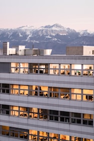 An office building with illuminated windows is pictured against a backdrop of snow-capped mountains during dusk. The warm glow from the interior contrasts with the cold hues of the mountain range in the background.