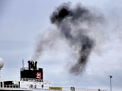 A ship's funnel releases a thick plume of black smoke into the overcast sky. Several people stand on the ship's deck, gazing at the smoke. The ship features a logo on the funnel and appears to be docked.