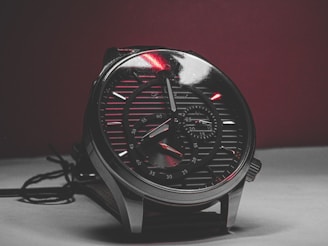 A close-up view of an analog wristwatch with a metallic finish. The watch face features multiple dials, including a chronograph, with intricate detailing. The surface is reflective, capturing light and adding a dynamic feel to the image.
