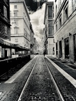 A black-and-white photograph of a narrow urban street lined with tall, old buildings. The street is paved with cobblestones and has tram tracks running down the center. Overcast skies are visible above, with a dramatic contrast between the clouds and the buildings. A lone person can be seen walking down the street in the distance.