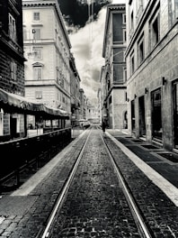 A black-and-white photograph of a narrow urban street lined with tall, old buildings. The street is paved with cobblestones and has tram tracks running down the center. Overcast skies are visible above, with a dramatic contrast between the clouds and the buildings. A lone person can be seen walking down the street in the distance.