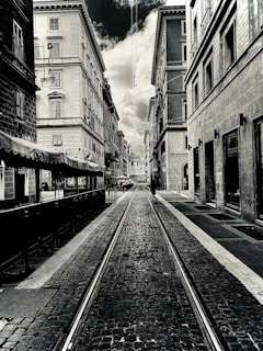 A black-and-white photograph of a narrow urban street lined with tall, old buildings. The street is paved with cobblestones and has tram tracks running down the center. Overcast skies are visible above, with a dramatic contrast between the clouds and the buildings. A lone person can be seen walking down the street in the distance.