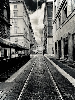 A black-and-white photograph of a narrow urban street lined with tall, old buildings. The street is paved with cobblestones and has tram tracks running down the center. Overcast skies are visible above, with a dramatic contrast between the clouds and the buildings. A lone person can be seen walking down the street in the distance.