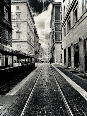 A black-and-white photograph of a narrow urban street lined with tall, old buildings. The street is paved with cobblestones and has tram tracks running down the center. Overcast skies are visible above, with a dramatic contrast between the clouds and the buildings. A lone person can be seen walking down the street in the distance.