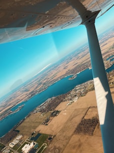 An aerial view of a landscape featuring a large body of water surrounded by fields and scattered buildings. The wing of an aircraft is visible in the foreground, indicating the photo was taken from within a plane. The sky is clear and blue, suggesting fair weather.