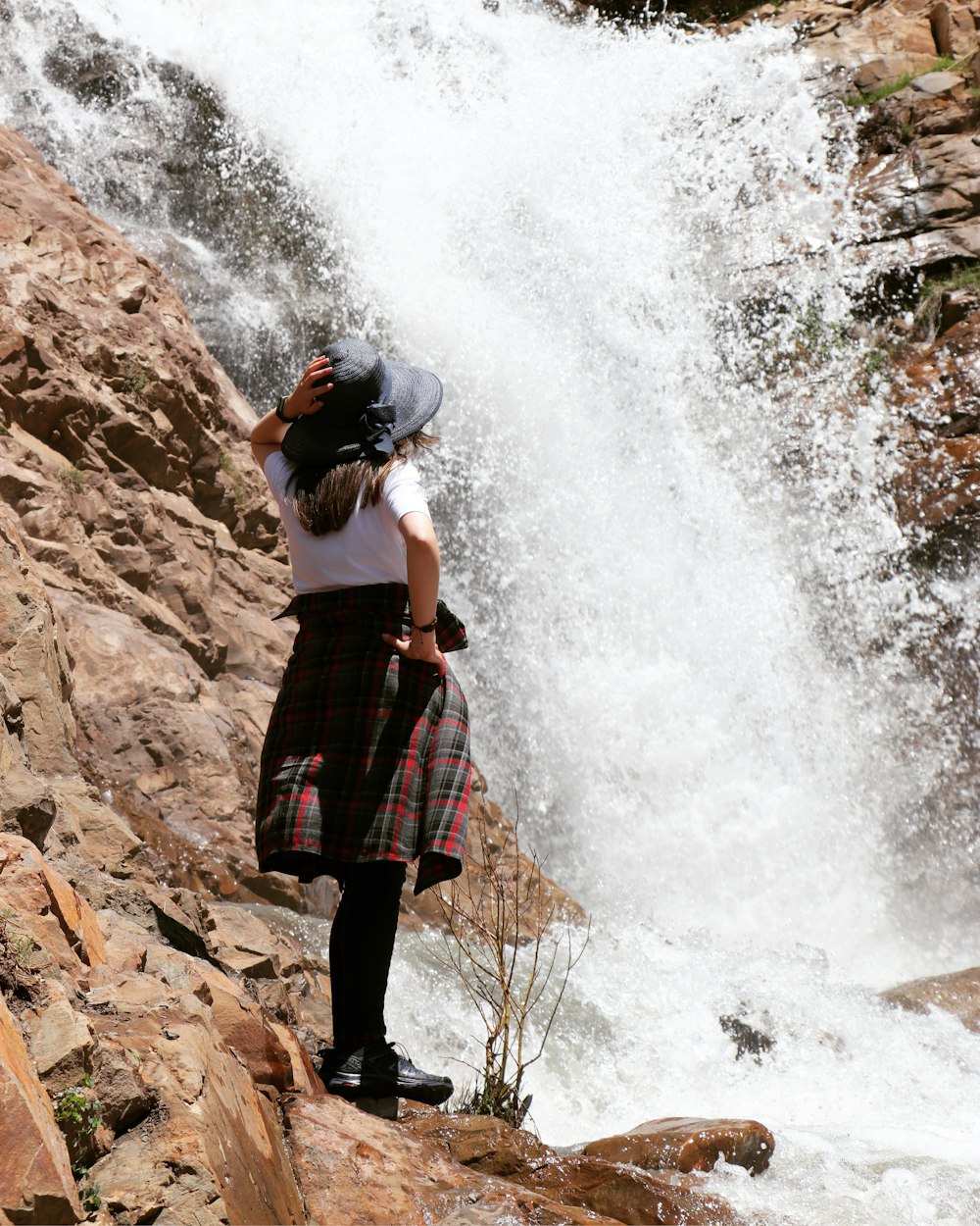 woman in black and red plaid skirt standing on brown rock near waterfalls during daytime