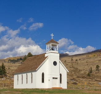 A small, white wooden church with a steeple and cross sits in a rural landscape. The building features arched windows and a dark round window near the top. It is surrounded by a grassy field, with a backdrop of hills and trees under a blue sky with scattered clouds.