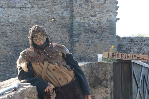 A person dressed in medieval fantasy attire, wearing a mask and chainmail, stands against a stone wall. They are leaning casually on a rock surface beside a wooden sign with 'KHORINIS' written on it. The background consists of rustic stone architecture.