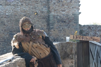 A person dressed in medieval fantasy attire, wearing a mask and chainmail, stands against a stone wall. They are leaning casually on a rock surface beside a wooden sign with 'KHORINIS' written on it. The background consists of rustic stone architecture.