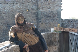 A person dressed in medieval fantasy attire, wearing a mask and chainmail, stands against a stone wall. They are leaning casually on a rock surface beside a wooden sign with 'KHORINIS' written on it. The background consists of rustic stone architecture.