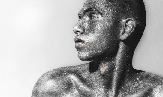 A person is covered in silver glitter, creating a metallic and shimmering effect against the skin. The face is turned in profile, revealing a strong jawline and an expression of contemplation or focus. The background is neutral, enhancing the sparkle and texture of the glitter.