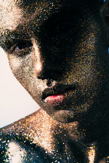 A close-up portrait of a person with their face and body covered in multicolored glitter, capturing light with a dramatic contrast between shadow and illumination. The glitter creates a textured, almost otherworldly skin effect with a play of colors reflecting under the light.