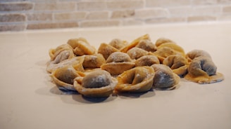 A group of uncooked dumplings arranged in a circular pattern on a light-colored surface. The background features a tiled wall with a beige brick pattern.
